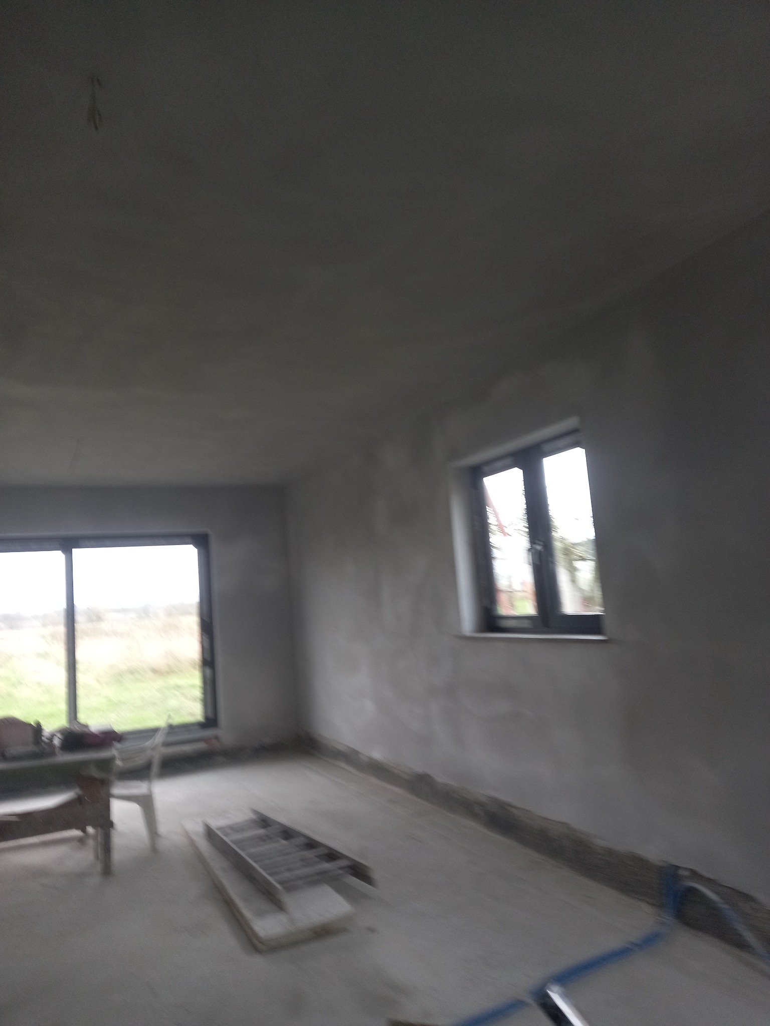 Wnętrze w trakcie prac wykończeniowych: surowe ściany z widocznymi nierównościami, okno z ciemną ramą, drabina aluminiowa leżąca na podłodze, oraz fragment instalacji w kolorze niebieskim.