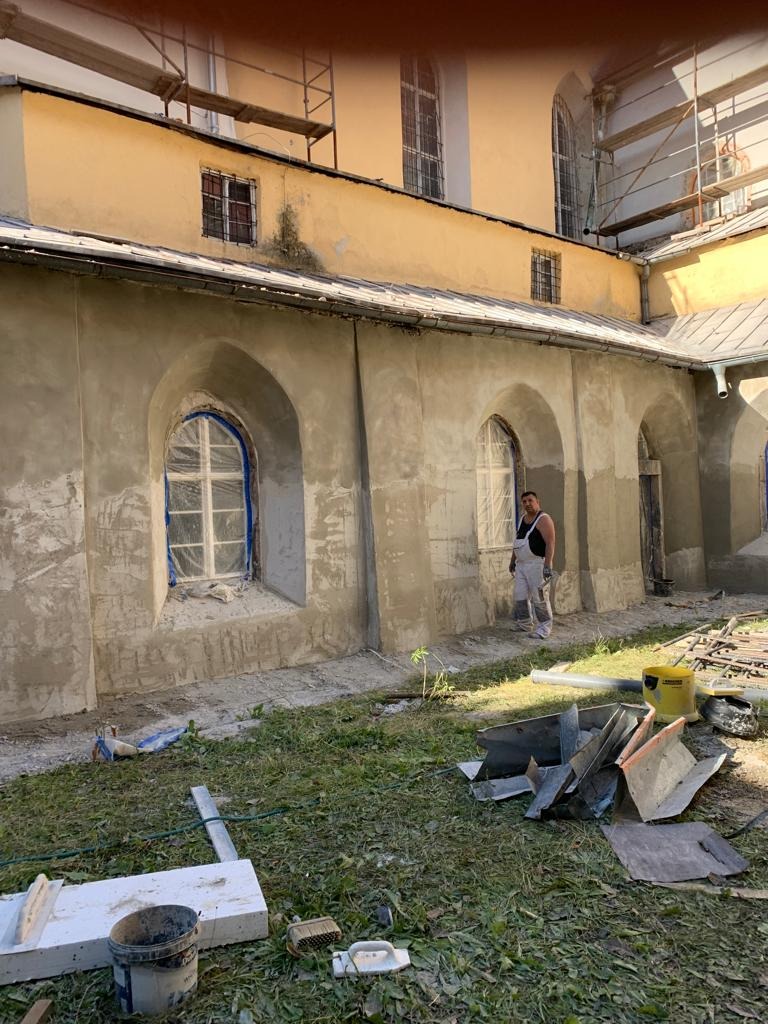 Prace na elewacji zabytkowego budynku z gotyckimi oknami, fragment ściany pokryty świeżym tynkiem, widoczne rusztowania i pracownik w kombinezonie, na trawie leżą narzędzia i odpady budowlane.