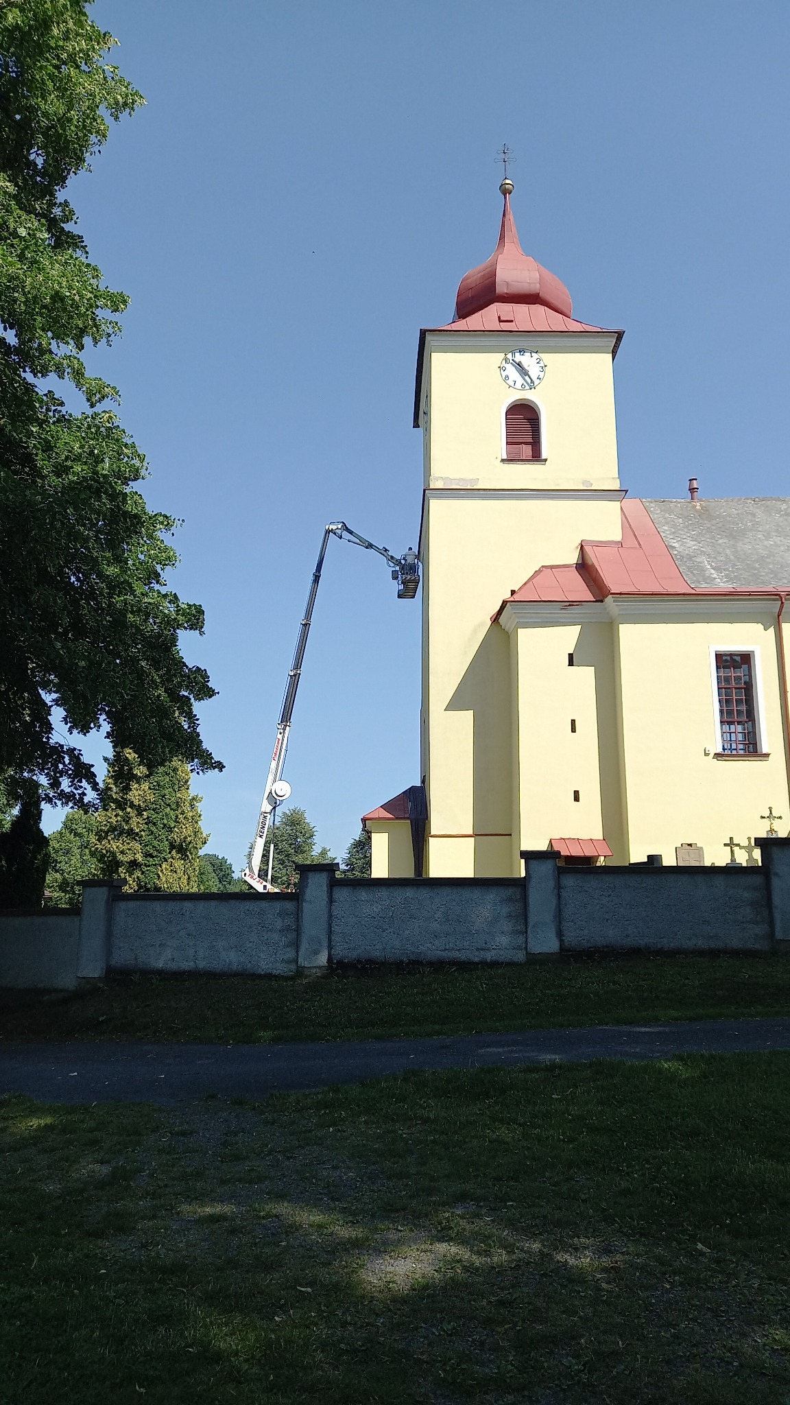 Żółty kościół z czerwoną dachówką, odnawiany z użyciem podnośnika koszowego na tle błękitnego nieba.