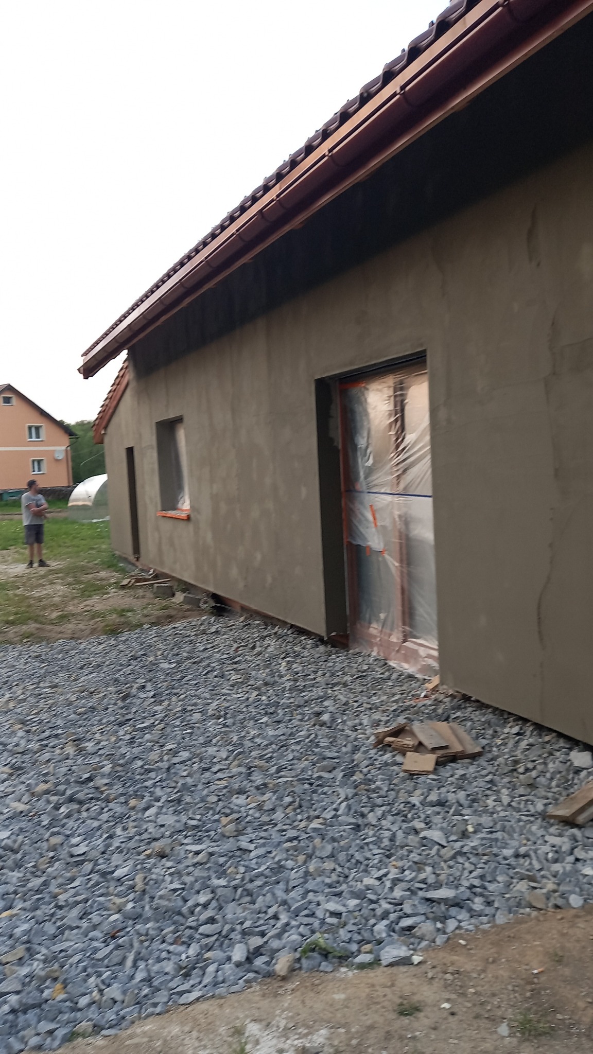 Świeżo otynkowana ściana budynku jednorodzinnego z zabezpieczonymi oknami folią, widok z boku na kamieniste podłoże i fragment trawnika.