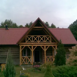 Dom z drewnianą werandą i nowym, czerwonym dachem z blachodachówki, na którym pracuje dekarz. Widoczna osoba w roboczym ubraniu na trawniku.