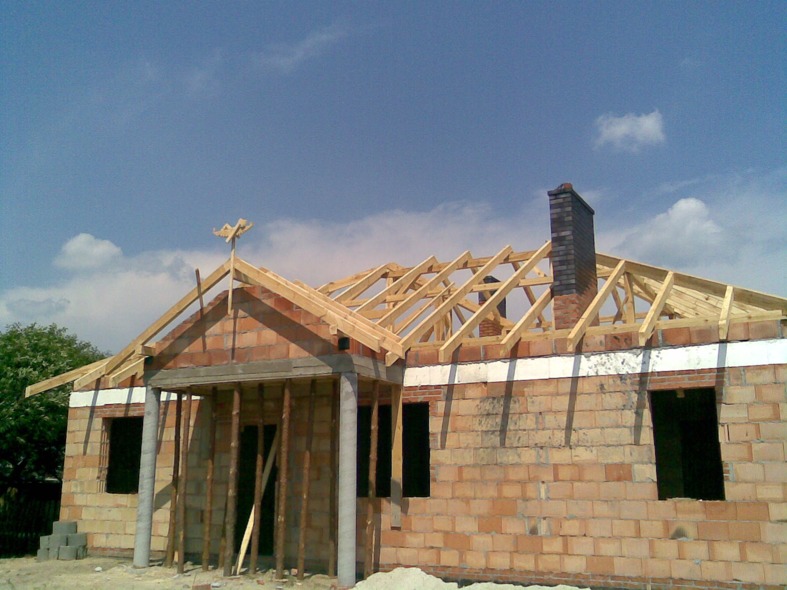 Surowy budynek z cegły z drewnianą więźbą dachową w trakcie montażu, widoczny komin i ozdobny wiatrowskaz na szczycie dachu.