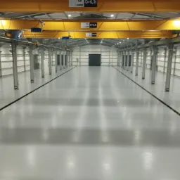 Jasnoszara, gładka podłoga w hali przemysłowej z widocznymi liniami dylatacyjnymi i metalowymi słupami podtrzymującymi konstrukcję dachu z suwnicą.