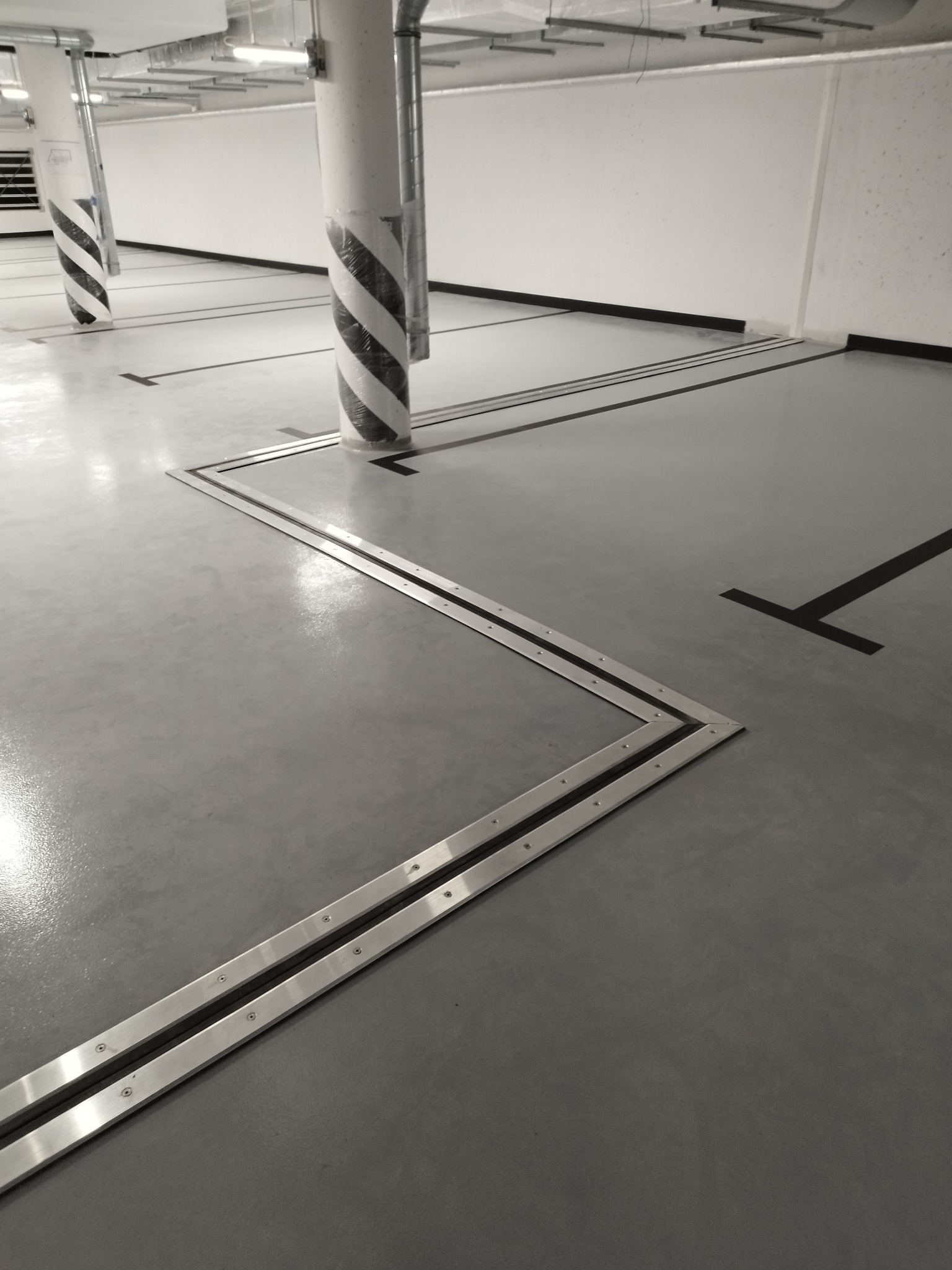 Szara posadzka w garażu podziemnym z metalowymi dylatacjami i czarnymi oznaczeniami miejsc parkingowych, słupy owinięte folią ochronną w biało-czarne pasy.