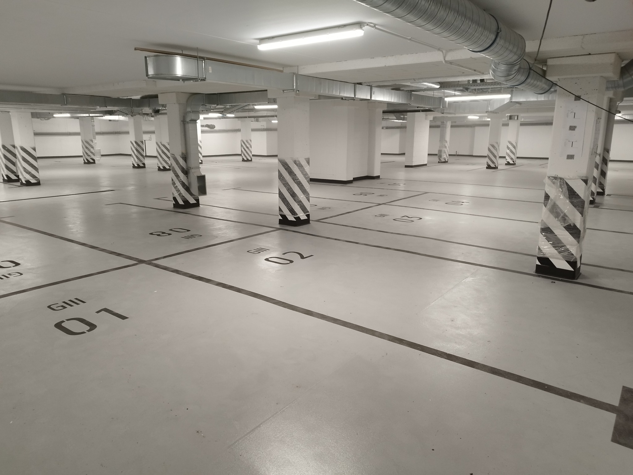 Jasnoszara posadzka w garażu podziemnym z czarnymi liniami wyznaczającymi miejsca parkingowe i numeracją, kolumny w biało-czarne pasy, oświetlenie sufitowe.