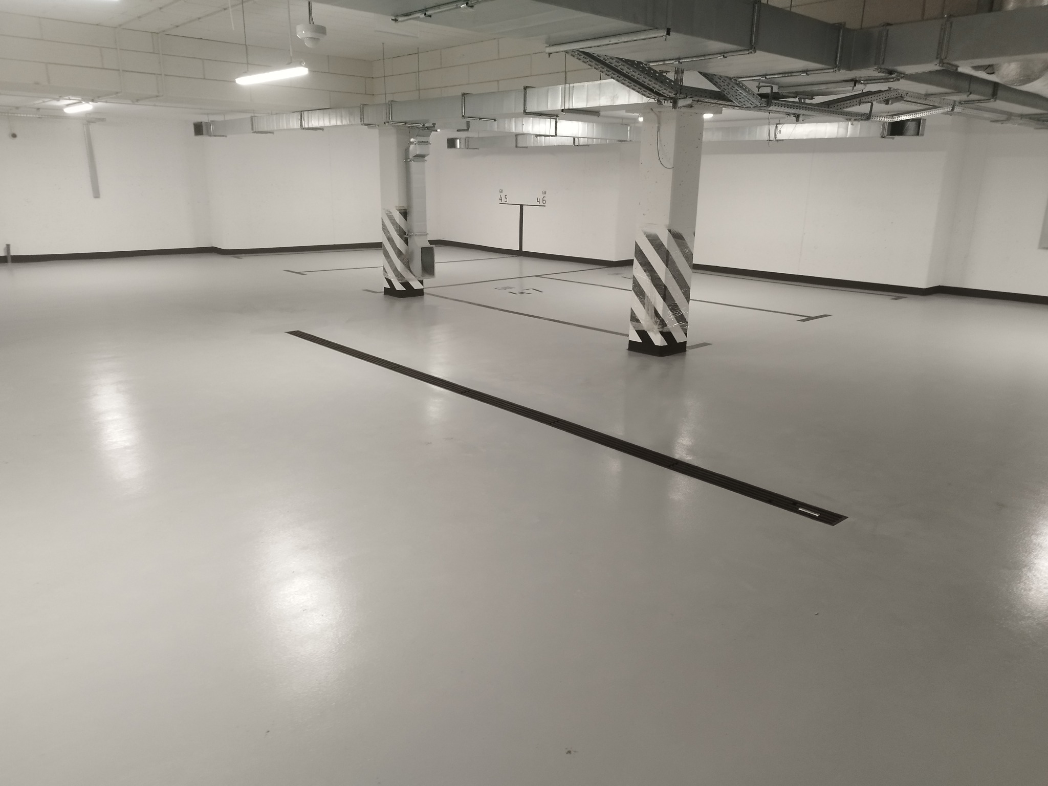 Jasnoszara, gładka posadzka w hali garażowej z oznaczonymi miejscami parkingowymi i słupami w biało-czarne pasy, oświetlona lampami sufitowymi i z widoczną instalacją wentylacyjną pod sufitem.