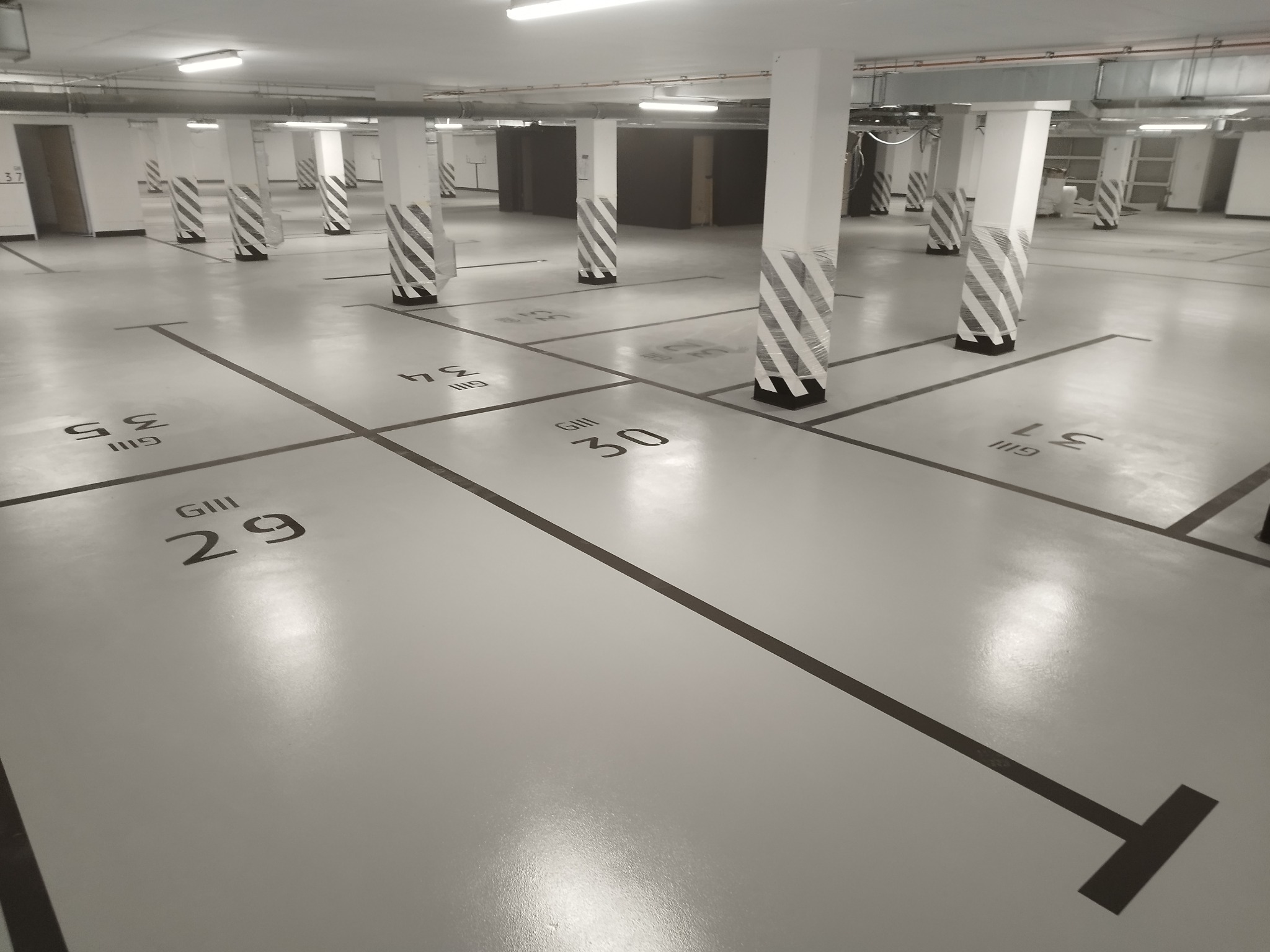 Jasna, pusta hala garażowa z szarą posadzką, czarnymi liniami wyznaczającymi miejsca parkingowe oznaczone numerami 'GIII 29', 'GIII 30', 'GIII 34', 'GIII 35' i 'GIII 37', z białymi kolumnami...