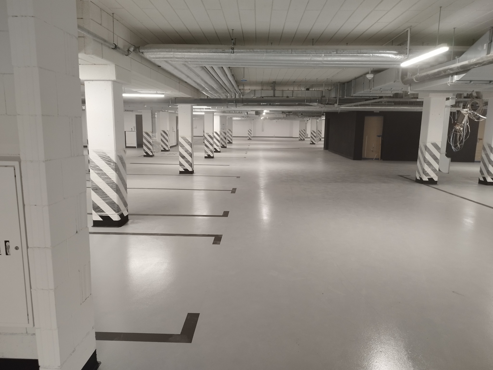 Pusta hala garażowa z szarą, jednolitą posadzką, kolumnami w biało-czarne pasy i widocznymi instalacjami pod sufitem.