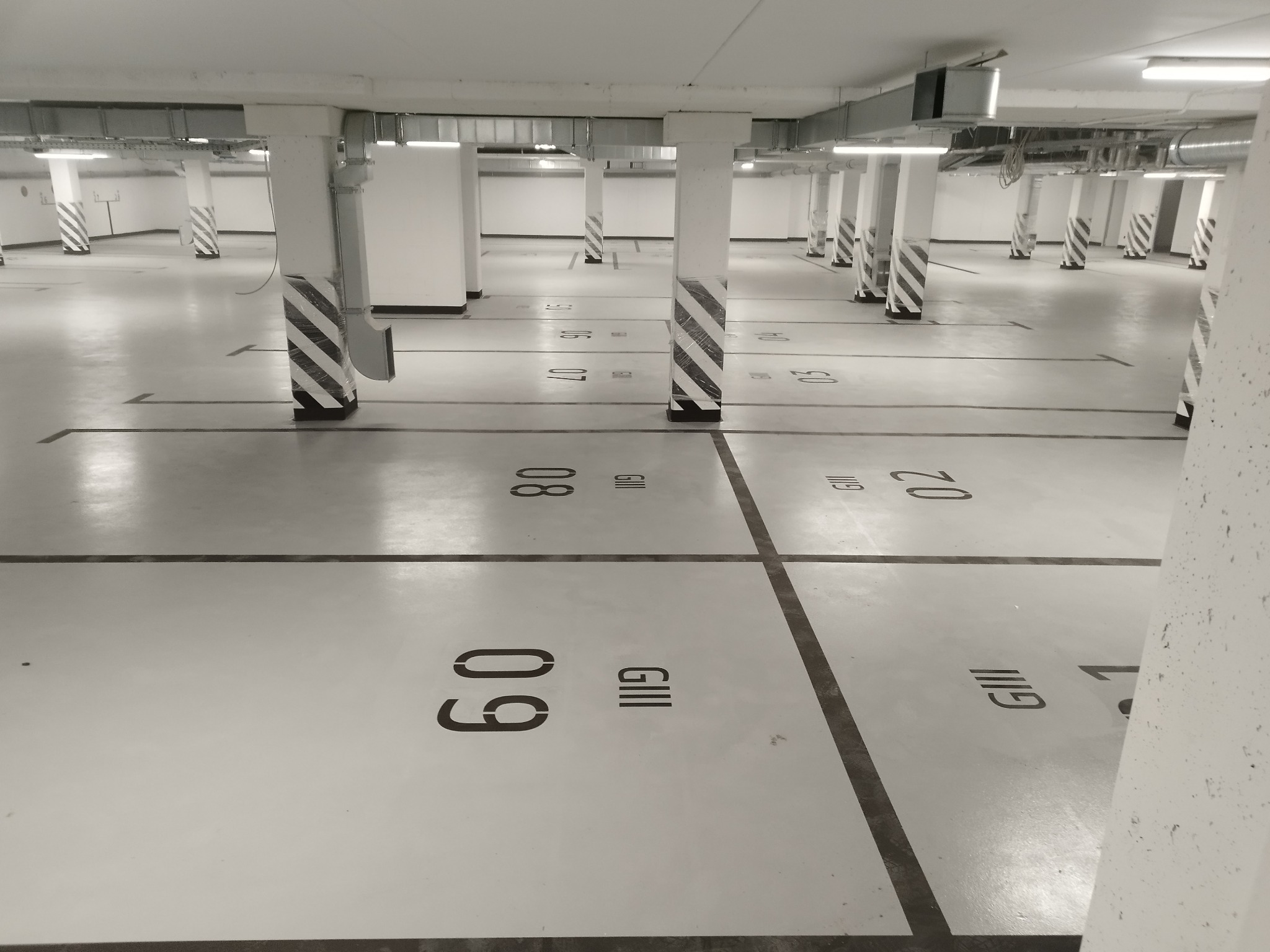 Jasna, pusta hala garażowa z szarą posadzką, czarnymi liniami wyznaczającymi miejsca parkingowe i numeracją stanowisk na posadzce; filary w biało-czarne pasy.