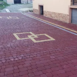 Ułożona, czerwona kostka brukowa z żółtymi akcentami w kształcie połączonych kwadratów i obrzeżami, widoczne odwodnienie liniowe przy budynku.