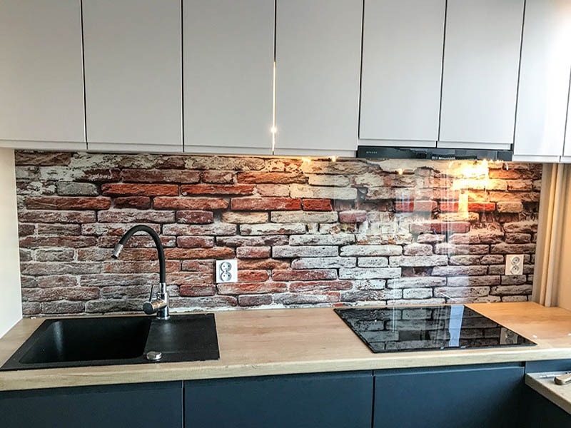 Szklany panel ścienny z motywem cegieł w kuchni, zamontowany nad blatem z czarnym zlewem i płytą indukcyjną, białe szafki wiszące.