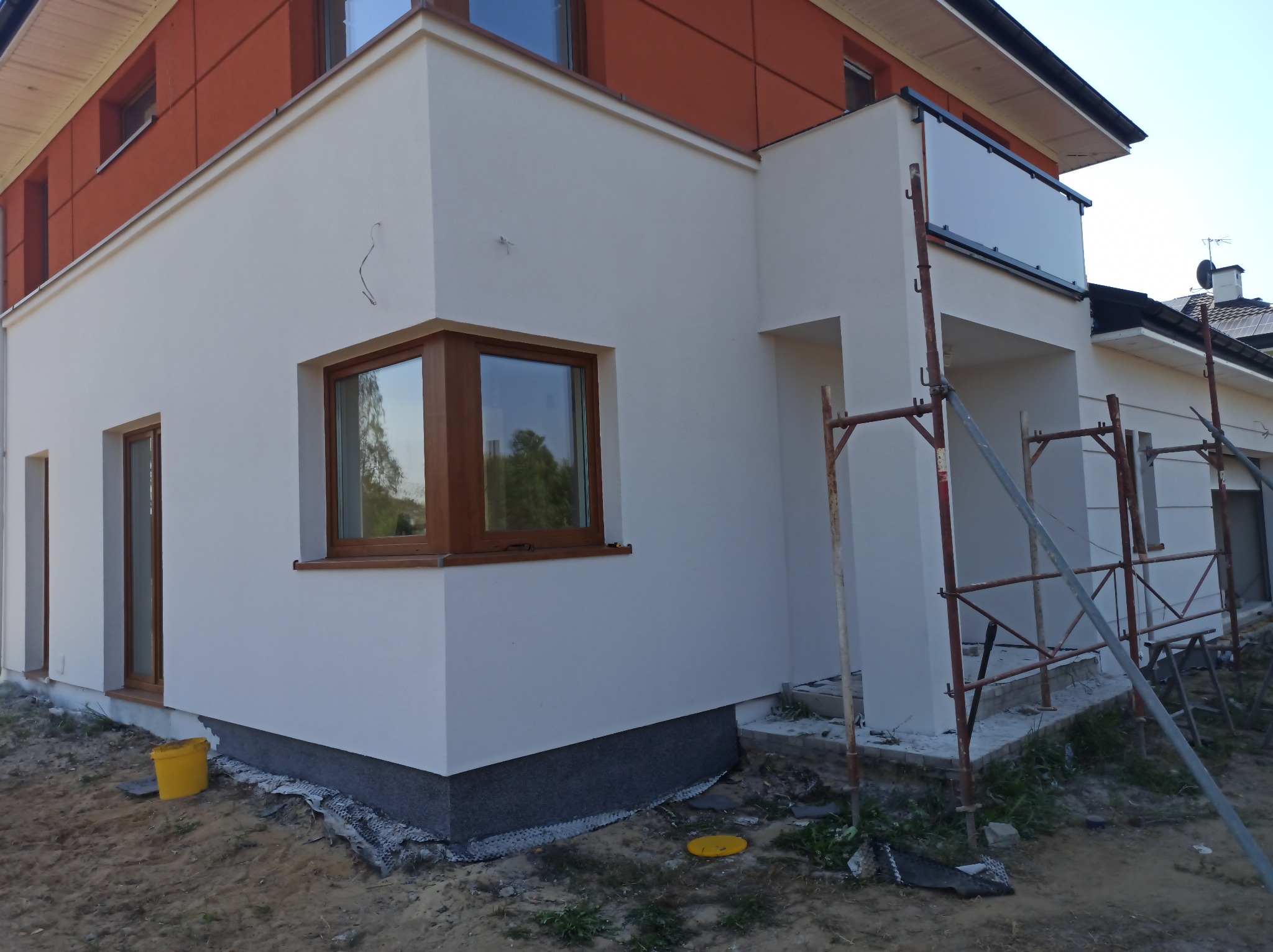 Nowoczesny dom w trakcie wykończenia elewacji. Widoczne okna z drewnianymi ramami, rusztowania i wiadro. Elementy w kolorze białym i pomarańczowym.
