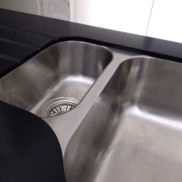 MEBLE XY NORBERT BOGDANOW - Dwukomorowy, stalowy zlew wpuszczony w czarny blat z ociekaczem. Widoczny fragment ściany z białymi płytkami. Nowoczesny design i precyzyjne wykonanie.