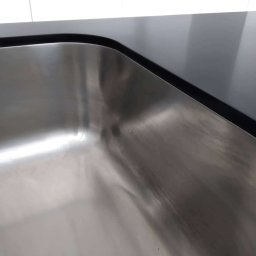 MEBLE XY NORBERT BOGDANOW - Detal zlewu ze stali nierdzewnej, wkomponowanego w czarny blat kuchenny z zaokrąglonym rogiem. Gładka powierzchnia, nowoczesny design, minimalistyczne ujęcie.