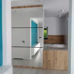 Nowoczesna zabudowa przedpokoju z lustrem, łącząca szafę i siedzisko w bieli i drewnie. Akcent turkusowej ściany dodaje świeżości wnętrzu.