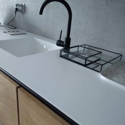 MEBLE XY NORBERT BOGDANOW - Nowoczesny blat kuchenny z białego kamienia z wbudowanym zlewozmywakiem i czarną armaturą. Widoczne drewniane fronty szafek i akcesoria kuchenne.