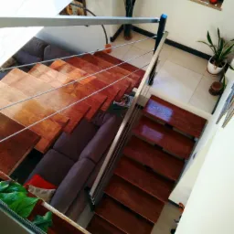 Drewniane schody z metalową balustradą, widok z góry na salon z szarą kanapą i roślinami w doniczkach na parapecie okiennym.