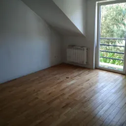 Pokój na poddaszu z wykończoną drewnianą podłogą, widok na zewnątrz przez okno balkonowe, biały grzejnik pod oknem, ściany w trakcie malowania.