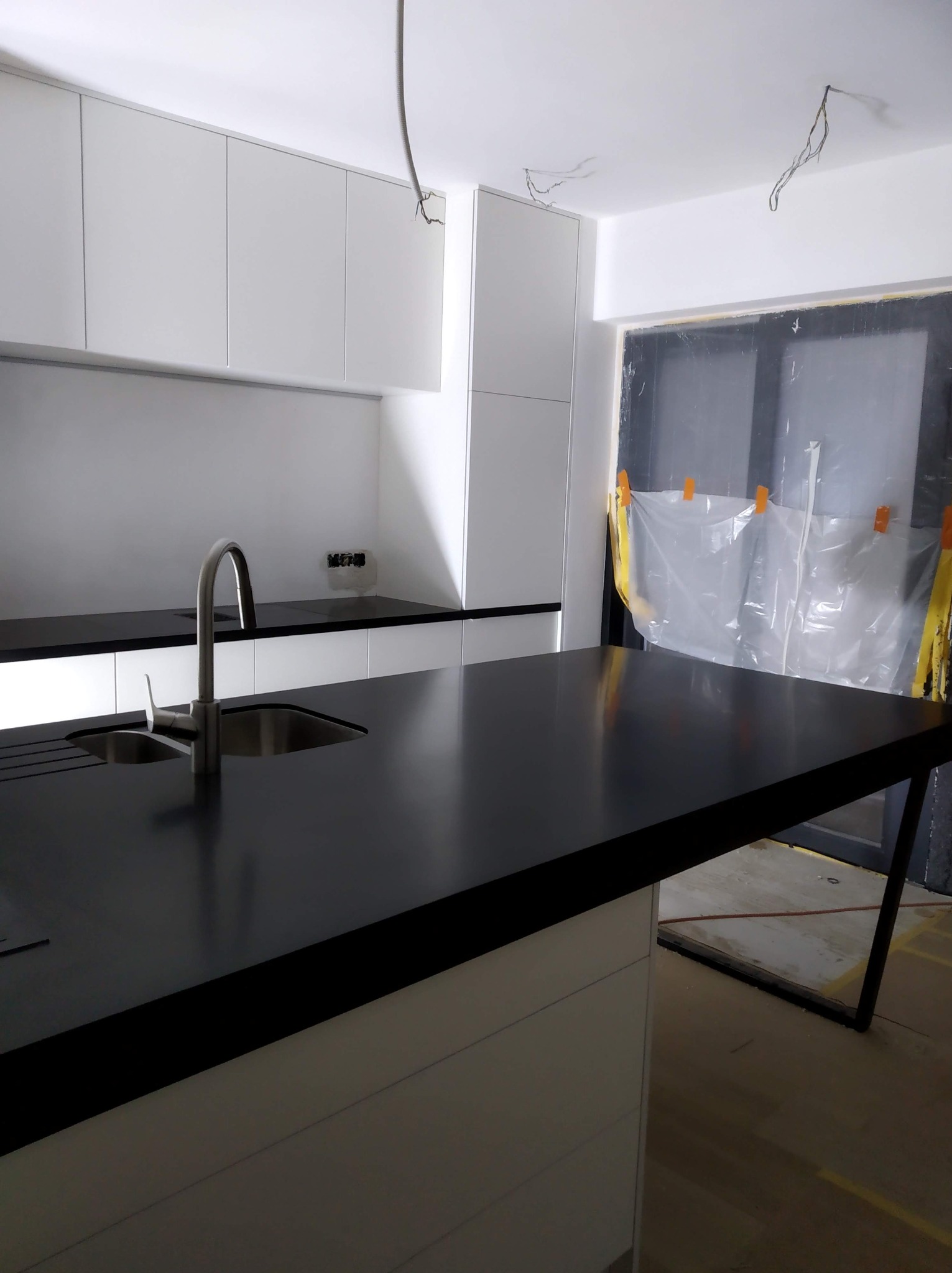 Nowoczesna kuchnia z czarnym blatem i białymi szafkami, widok na zlew i baterię. Okno zabezpieczone folią malarską. Minimalistyczny design, surowy stan wykończenia.