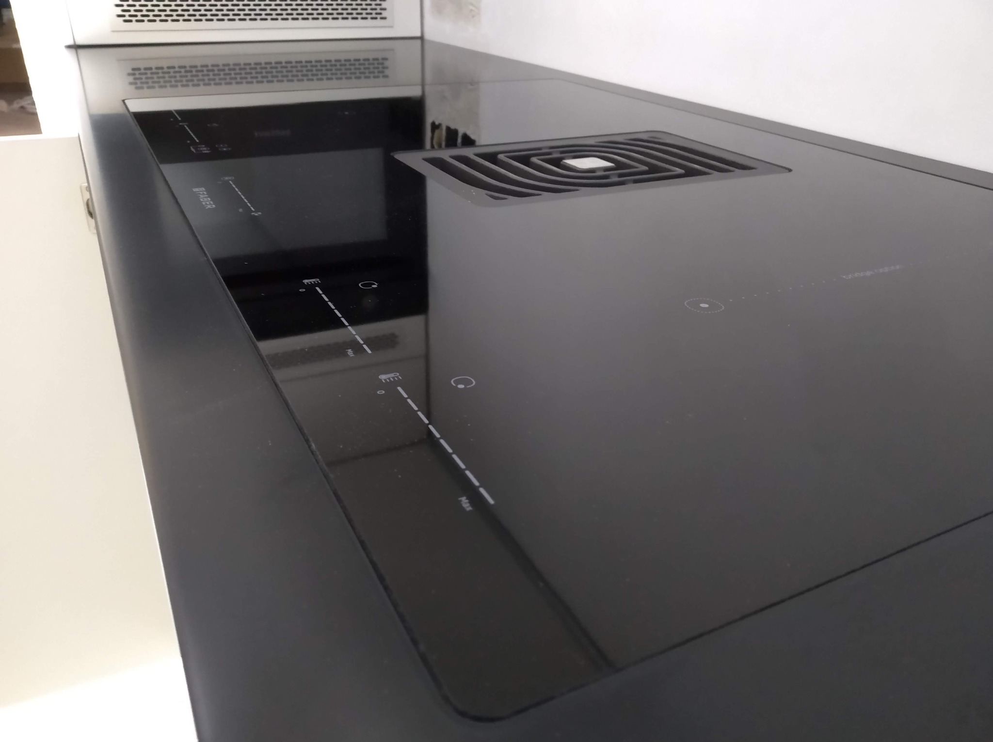 Czarna, błyszcząca płyta indukcyjna z wbudowanym okapem, marki Faber. Minimalistyczny design, widoczne panele sterowania dotykowego i kratka wentylacyjna. Nowoczesna kuchnia.