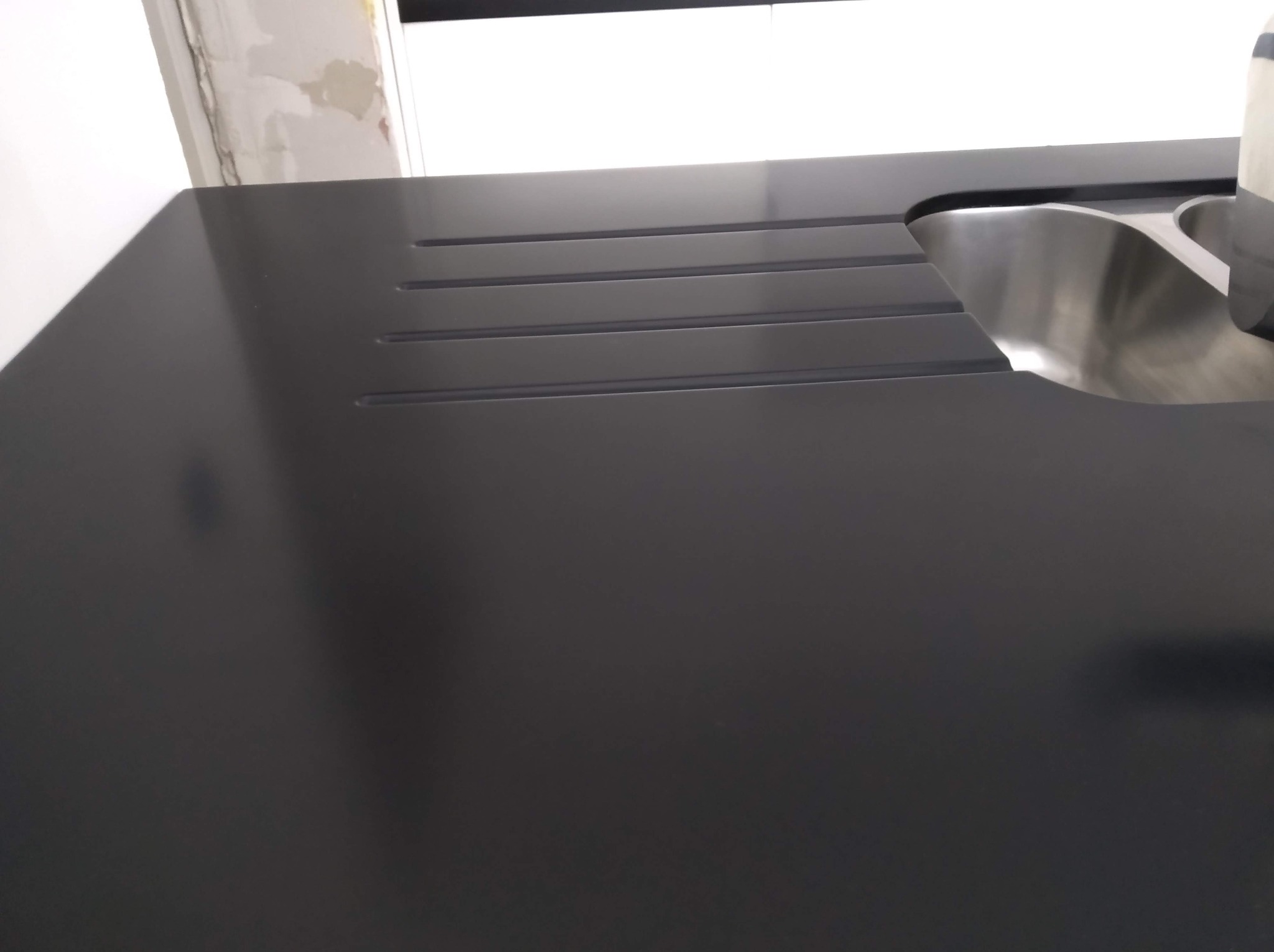 Czarny, matowy blat kuchenny z ociekaczem i zlewem ze stali nierdzewnej. Minimalistyczny design, widoczne niedoskonałości ściany w tle.