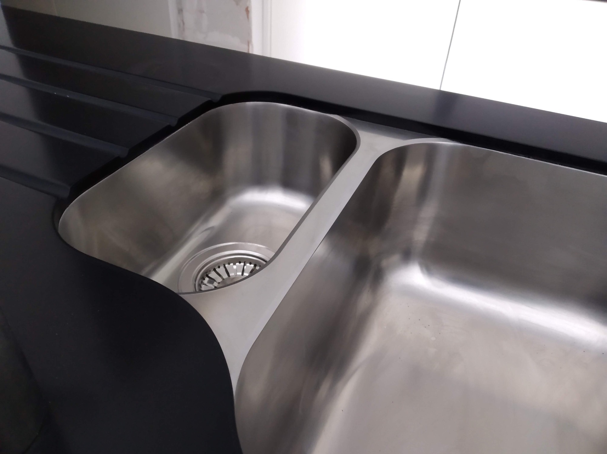 Dwukomorowy, stalowy zlew wkomponowany w czarny, matowy blat z ociekaczem. Minimalistyczny design, nowoczesne wykończenie, jasne oświetlenie.