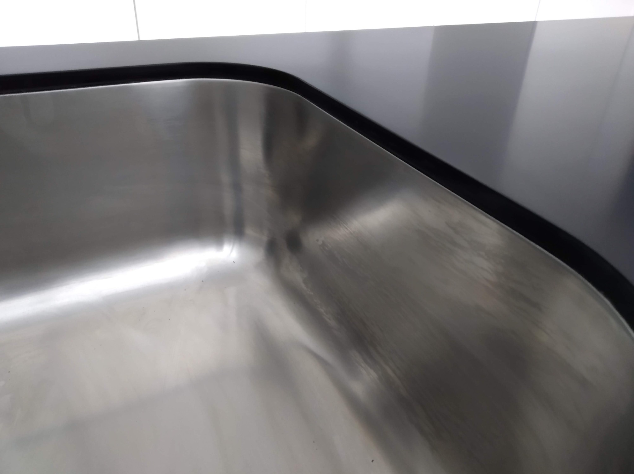 Detal zlewu ze stali nierdzewnej, wkomponowanego w czarny blat kuchenny. Widoczne białe płytki w tle, oświetlenie górne.