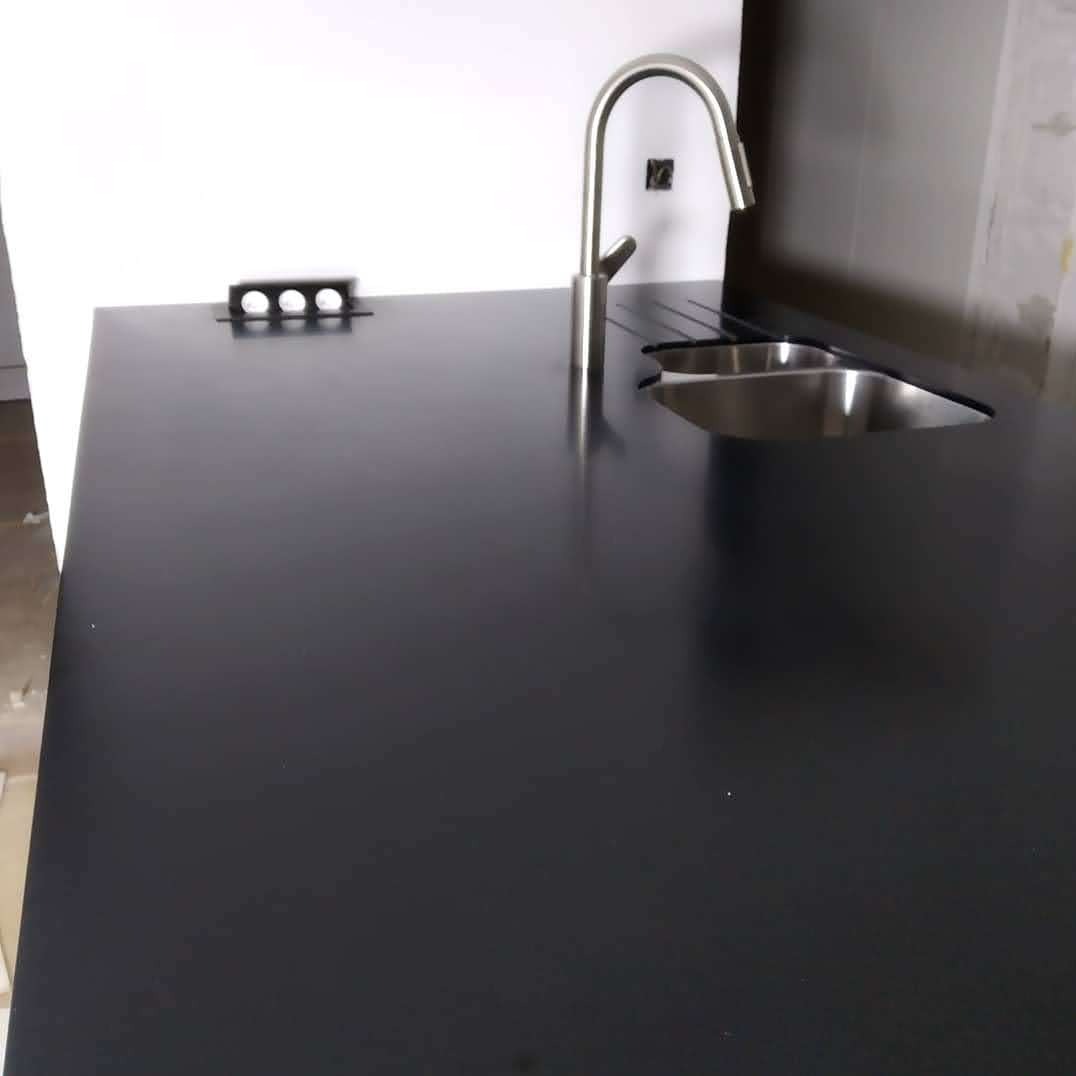 Czarny blat kuchenny z metalowym zlewem i baterią, minimalistyczny design, oświetlenie punktowe, nowoczesna estetyka wnętrza.