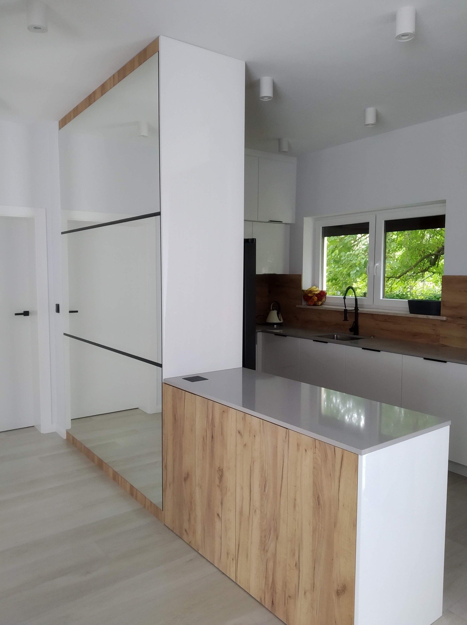 Nowoczesna kuchnia z wyspą w odcieniach bieli i drewna, z lustrzanym panelem oddzielającym przestrzeń. Minimalistyczne oświetlenie punktowe i duży, jasny pokój.