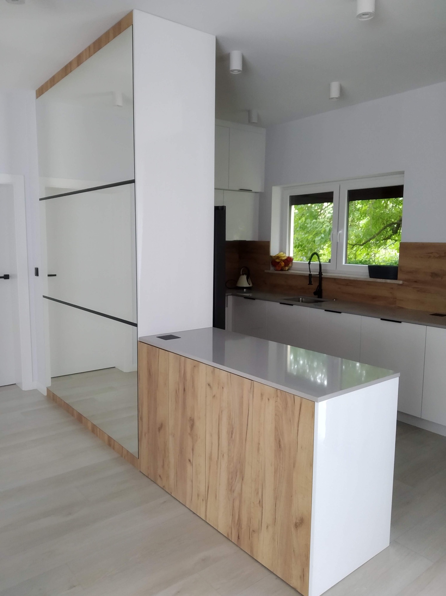 Nowoczesna kuchnia z wyspą i zabudową z lustrem. Blat szary, fronty białe i drewnopodobne. Widok na okno z zielenią. Minimalistyczny design.