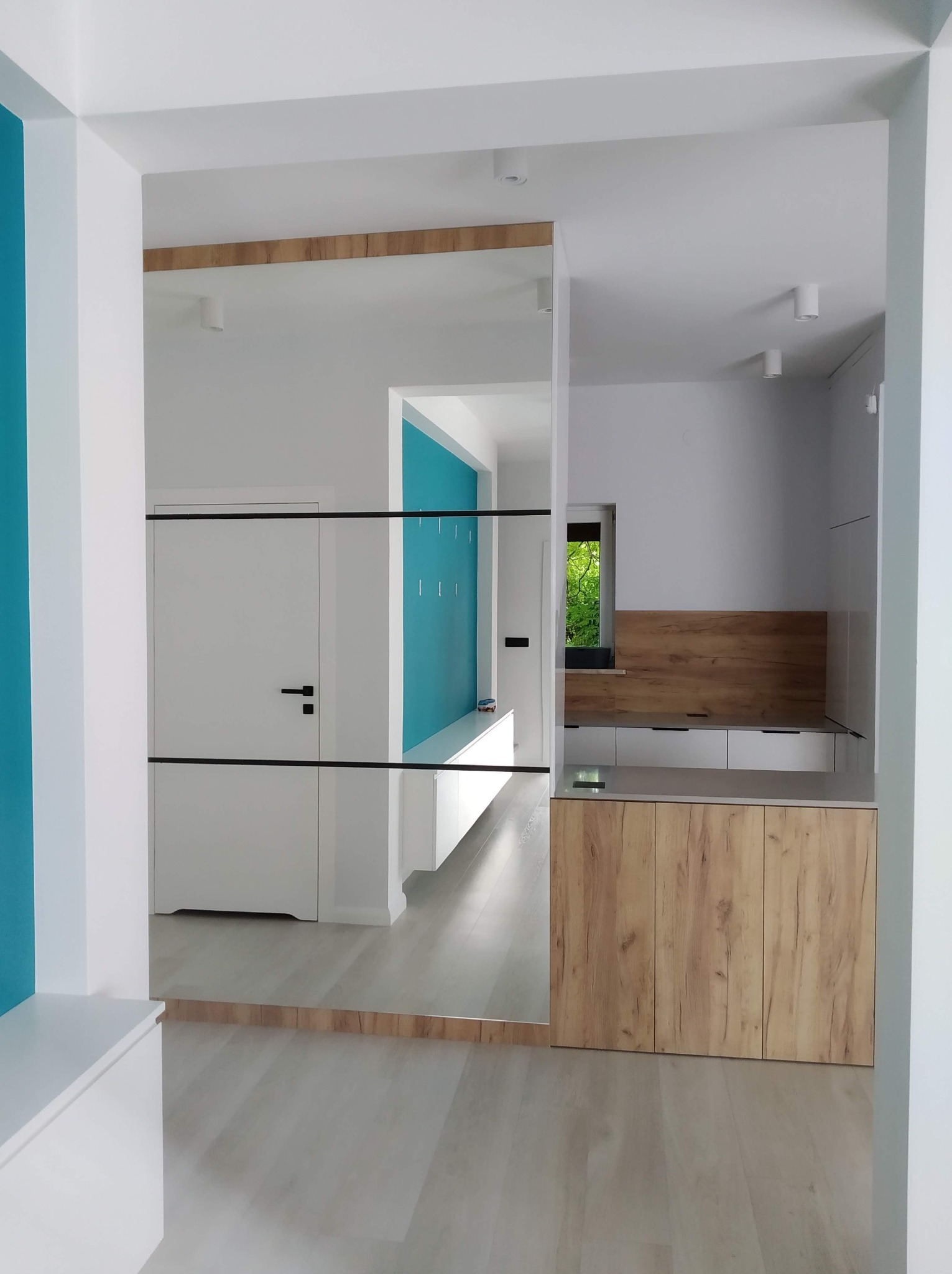 Nowoczesna zabudowa przedpokoju z lustrem, łącząca szafę i siedzisko w bieli i drewnie. Akcent turkusowej ściany dodaje świeżości wnętrzu.