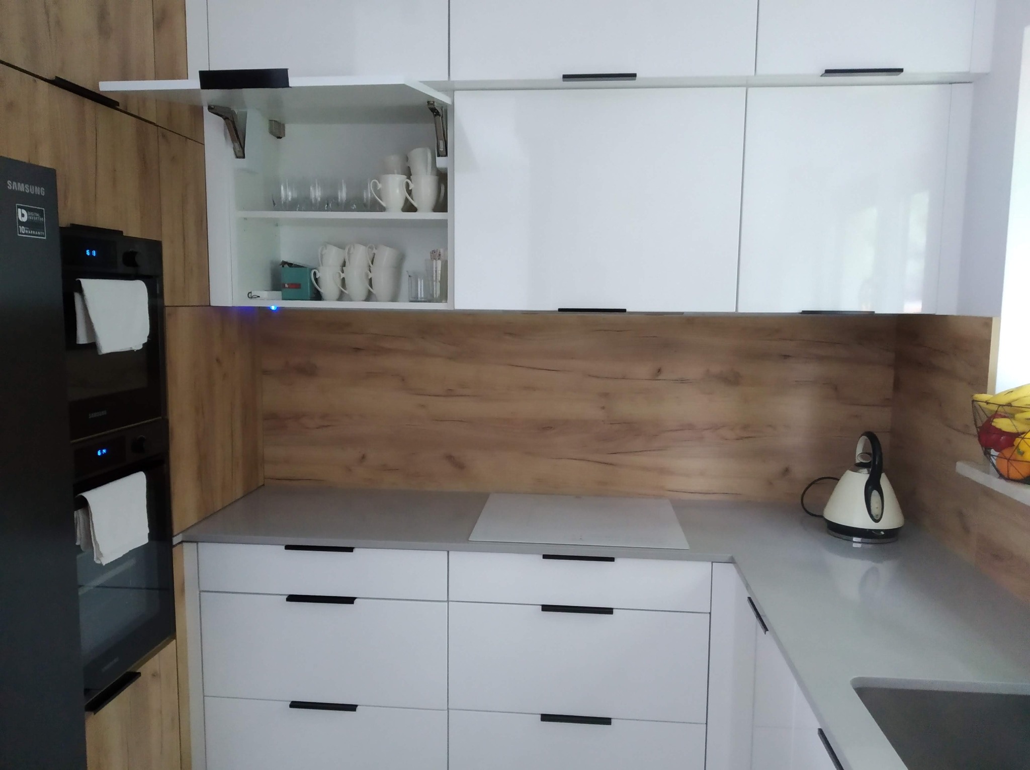 Nowoczesna kuchnia: białe fronty szafek z czarnymi uchwytami, drewnopodobny panel, otwarty górny szafka z naczyniami, czarny piekarnik Samsung.