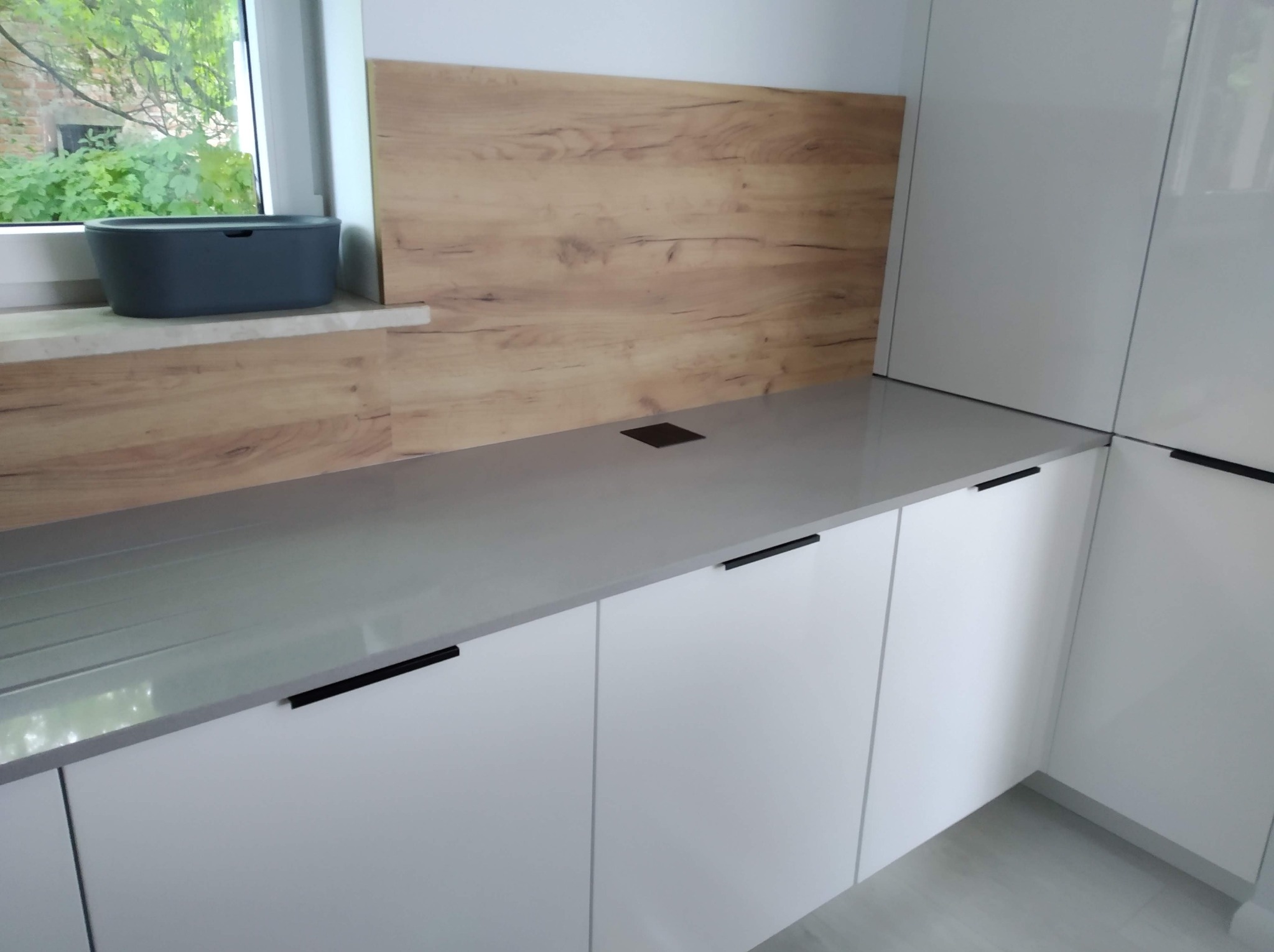 Nowoczesna zabudowa kuchenna z białymi, gładkimi frontami, szarym blatem i drewnopodobnym panelem nad blatem. Minimalistyczne, czarne uchwyty podkreślają linię szafek.