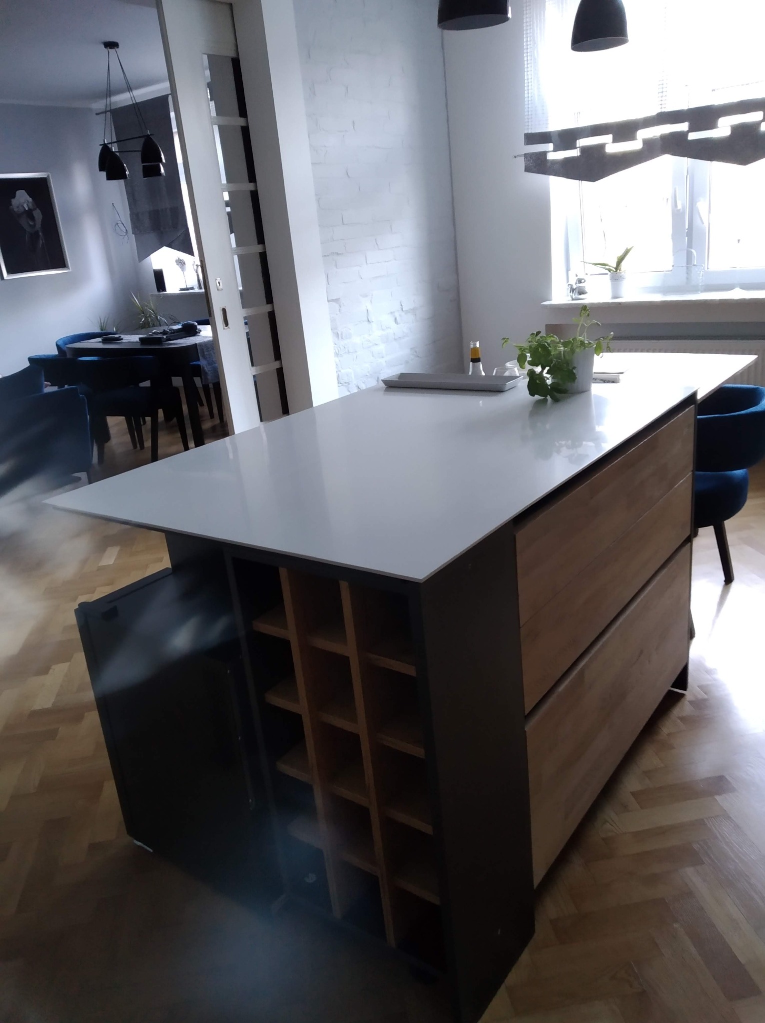 Nowoczesna wyspa kuchenna z białym blatem, drewnianymi szufladami i regałem na wino. W tle widoczna jadalnia i ceglana ściana.