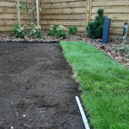 Połowa działki z przygotowaną ziemią, a druga połowa z rozłożoną świeżą trawą z rolki, widoczna listwa pomiarowa, drewniany płot w tle.