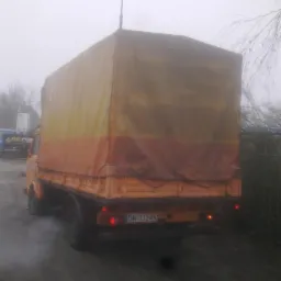 Żółto-pomarańczowy samochód dostawczy z plandeką, widok z tyłu, tablica rejestracyjna DW-3324N, lekka mgła w tle.