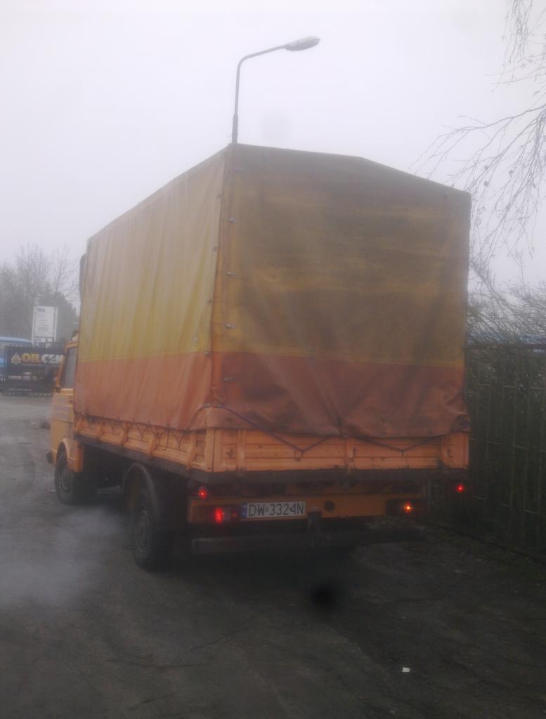 Żółto-pomarańczowy samochód dostawczy z plandeką, widok z tyłu, tablica rejestracyjna DW-3324N, lekka mgła w tle.