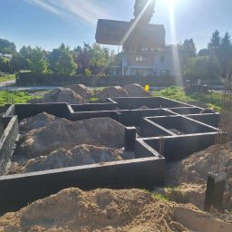 SZWAGIER-KOP SPÓŁKA Z OGRANICZONĄ ODPOWIEDZIALNOŚCIĄ - Fundamenty domu jednorodzinnego w trakcie zasypywania ziemią, widoczna zbrojona konstrukcja i koparka w tle.