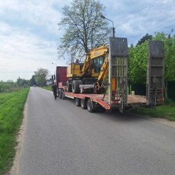 SZWAGIER-KOP SPÓŁKA Z OGRANICZONĄ ODPOWIEDZIALNOŚCIĄ - Transport niskopodwoziowy: żółta koparka kołowa na czerwonej platformie transportowej, zabezpieczona. Widok z tyłu na drogę z zielenią po bokach.