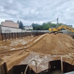 SZWAGIER-KOP SPÓŁKA Z OGRANICZONĄ ODPOWIEDZIALNOŚCIĄ - Żółta koparka Komatsu na hałdzie piasku w wykopie zabezpieczonym stalowymi ściankami Larsena, w tle budynki i pochmurne niebo. Prace ziemne w toku.