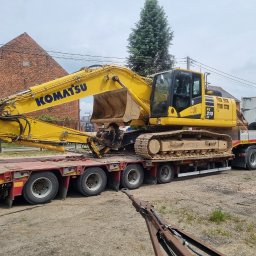 SZWAGIER-KOP SPÓŁKA Z OGRANICZONĄ ODPOWIEDZIALNOŚCIĄ - Żółta koparka Komatsu PC 210 LC, brudna od ziemi, transportowana na niskopodwoziowej przyczepie ciężarówki, z widocznym fragmentem haka holowniczego na pierwszym planie.