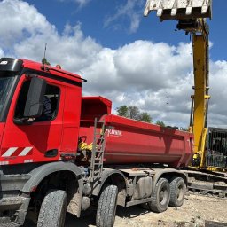 SZWAGIER-KOP SPÓŁKA Z OGRANICZONĄ ODPOWIEDZIALNOŚCIĄ - Czerwona ciężarówka i koparka w trakcie prac ziemnych na placu budowy, z widocznymi elementami infrastruktury i błękitnym niebem w tle.
