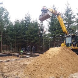 SZWAGIER-KOP SPÓŁKA Z OGRANICZONĄ ODPOWIEDZIALNOŚCIĄ - Żółta koparka z łyżką nad fundamentem domu, grupa robotników zagęszcza piasek w lesie. Stos piasku na pierwszym planie.