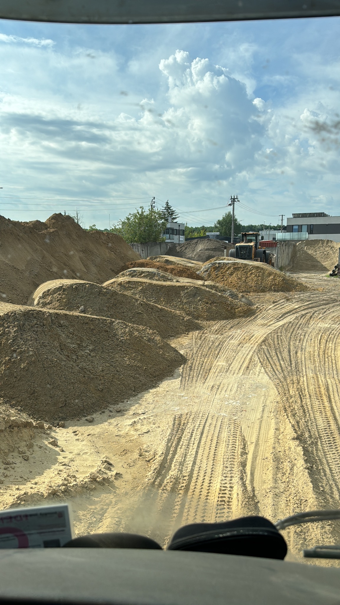 Kopalnia piasku z hałdami i śladami opon ciężkiego sprzętu. W tle budynki i błękitne niebo z chmurami, widok z kabiny pojazdu.