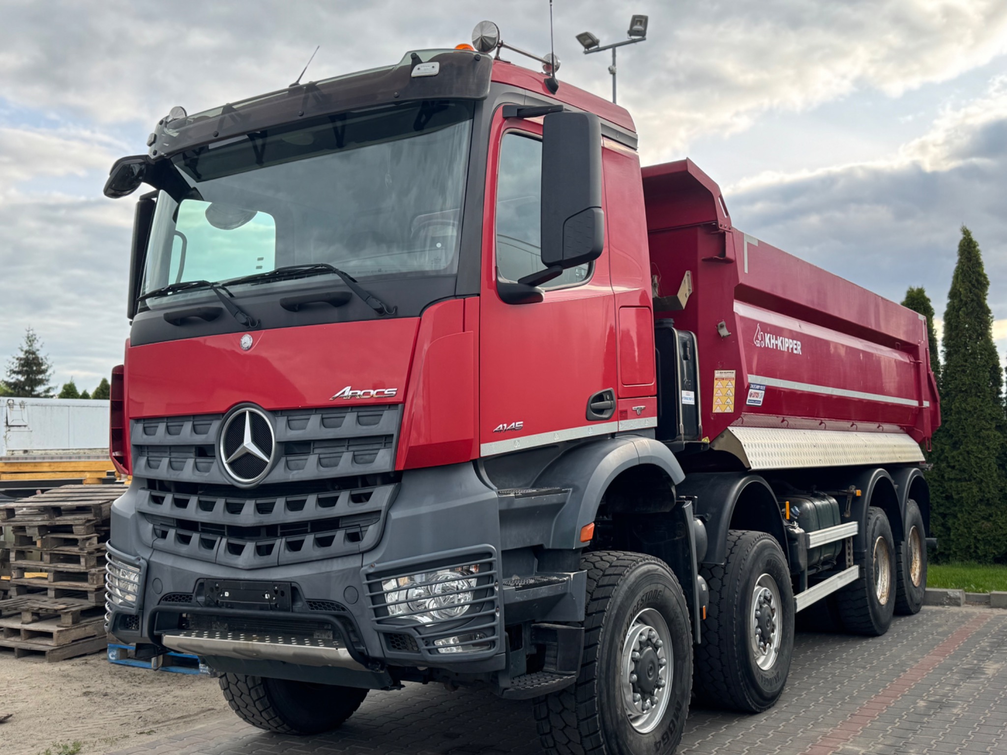 Czerwona wywrotka Mercedes-Benz Arocs 4145 z logo KH-Kipper, zaparkowana na kostce brukowej, z paletami w tle i pochmurnym niebem.