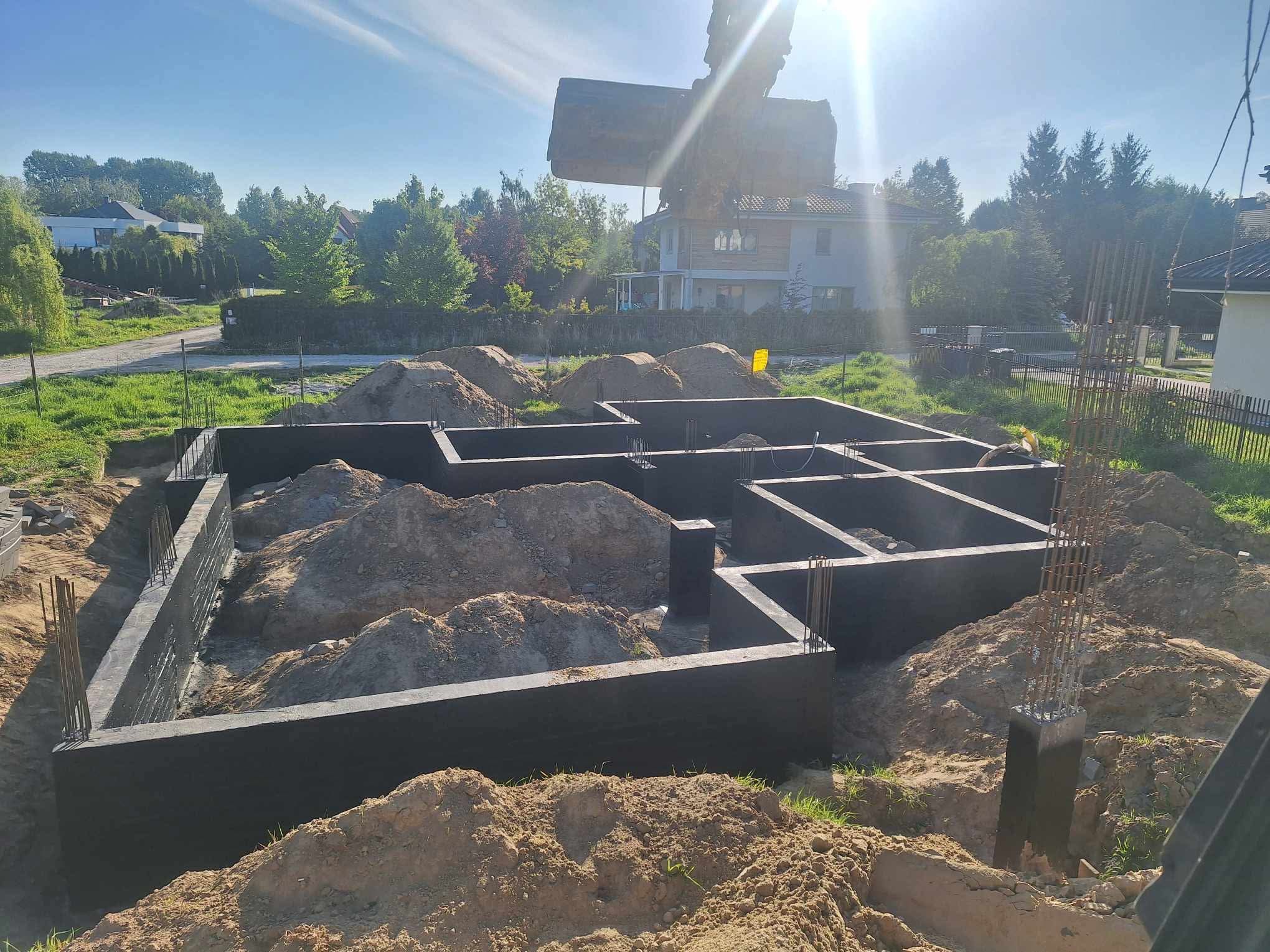 Fundamenty domu jednorodzinnego w trakcie zasypywania ziemią, widoczna zbrojona konstrukcja i koparka w tle.