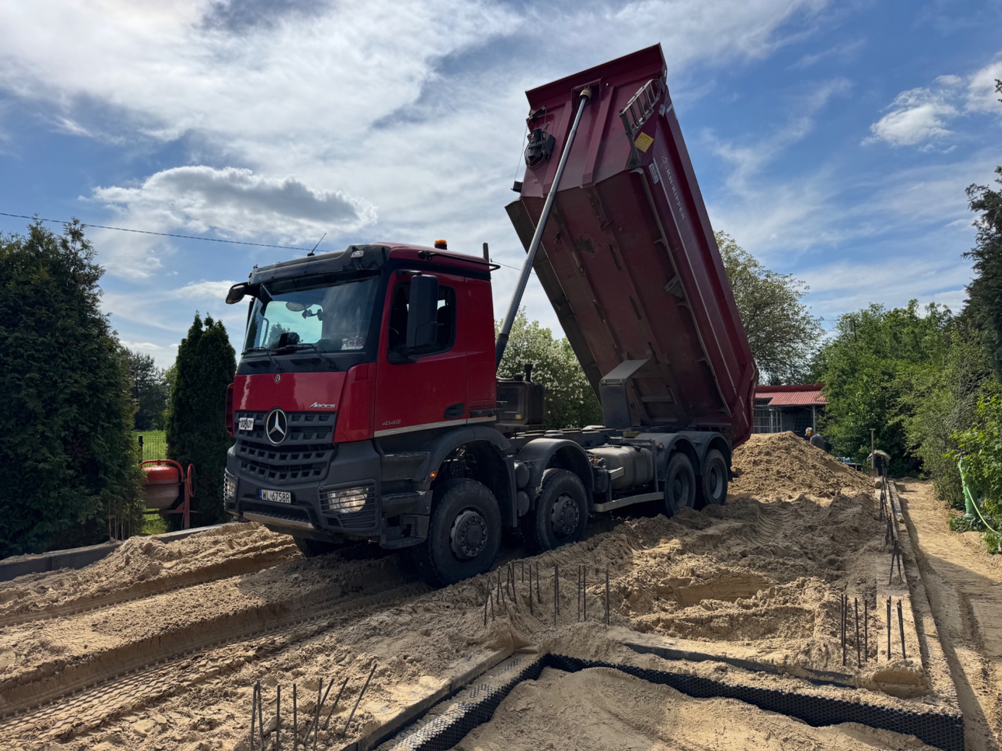 Czerwona wywrotka Mercedes Aroc w trakcie rozładunku piasku na placu budowy z widocznymi elementami szalunku i zbrojenia pod fundamenty.