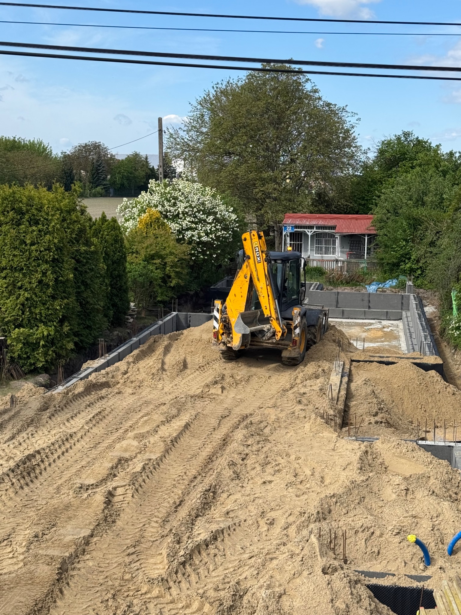 Żółta koparko-ładowarka na placu budowy, wyrównuje piasek w wykopie pod fundamenty. Widoczne ślady opon i częściowo zalane fundamenty.