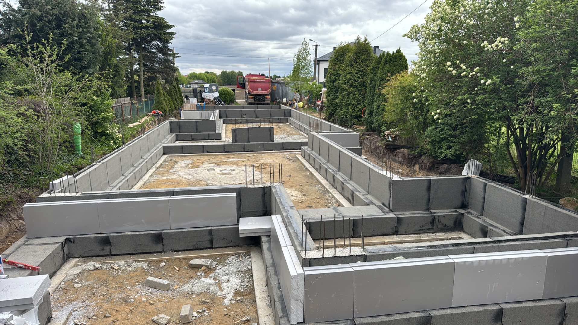 Fundamenty domu z szalunków systemowych, częściowo wypełnione piaskiem. Na posesji stoi czerwona wywrotka. Widok na zieleń i niebo.