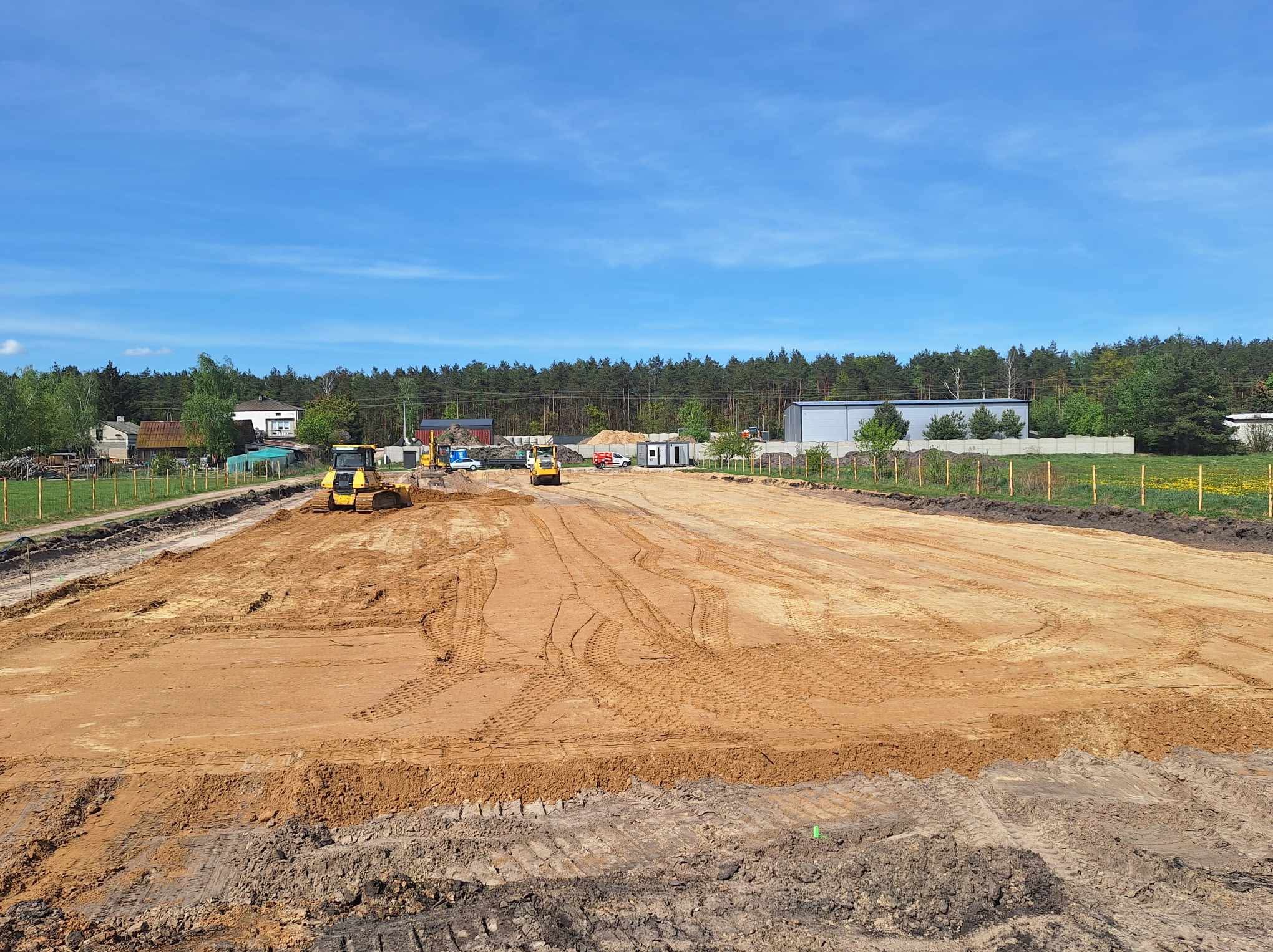 Rozległy teren wyrównywany przez dwie żółte spycharki w słoneczny dzień. Ślady opon na piasku, w tle budynki i las. Widok z poziomu gruntu.
